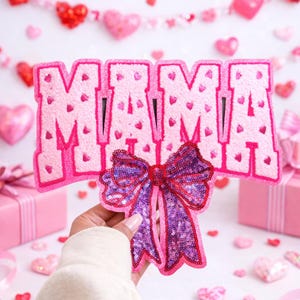 May include: A pink felt applique spelling "MAMA" with small heart details. Below, a sequined purple bow with red trim is held in front of the letters. The background features pink hearts and gift boxes, creating a festive, Valentine's Day theme.