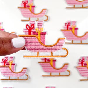 May include: Pink and white embroidered Christmas sleigh with red and yellow presents. The sleigh is decorated with gold trim and has a white background.