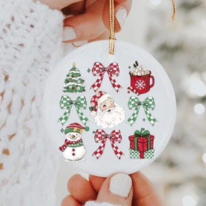 May include: A white ceramic Christmas ornament with a gold string, featuring festive illustrations. The design includes a Christmas tree, bows, Santa Claus, a mug of hot cocoa, a snowman, and a wrapped gift.