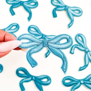 May include: Embroidered light blue bow patches on a white background. The bows are detailed with a satin stitch and have a classic bow shape. These patches are suitable for various craft projects.