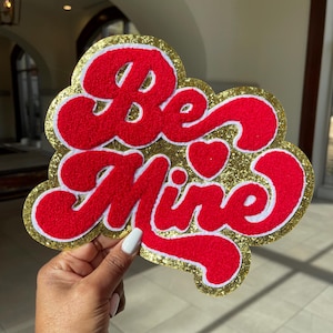 May include: A red and gold glitter felt applique with the words "Be Mine" in a retro font. A small heart is included in the design.
