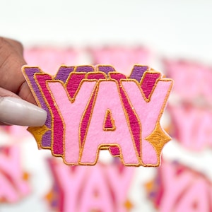 May include: Pink and purple embroidered patch with the word "YAY" in a bubble letter style. The patch has a gold border and a starburst design. The patch is held in a hand.