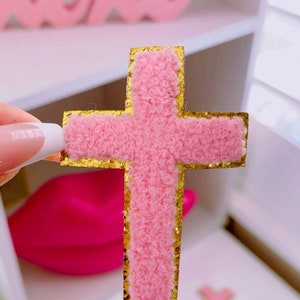 May include: A pink cross with a gold glitter outline. The cross is made of a fluffy material.