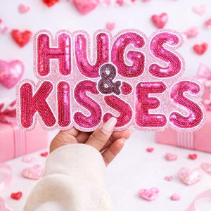May include: A pink sequined sign that reads "HUGS & KISSES" is held in front of a white background decorated with pink and red hearts. The sign is in a bubble letter style with a white border. Pink gift boxes and ribbons are also visible.