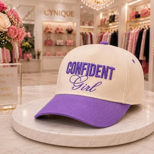 May include: A baseball cap with a cream-colored crown and a purple brim. The front features the text "CONFIDENT" in bold purple block letters above the word "Girl" in a purple cursive script. The cap has a purple button on the top center.