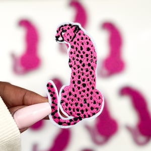 May include: Pink cheetah iron-on patch with black spots. The patch is embroidered and has a white outline.