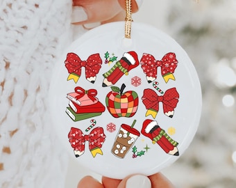 May include: Round white ceramic Christmas ornament with a gold hanging string. The ornament is decorated with colourful illustrations of bows, pencils, books, an apple, snowflakes, and a coffee drink.