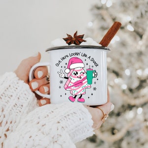 May include: White enamel mug with a black rim and handle. The mug features a cartoon Christmas tree wearing a Santa hat and holding a green drink. The text on the mug reads "Out Here Lookin' Like A Snack".