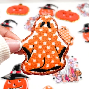 May include: Orange and white checkered ghost patch with black eyes. The patch is made of fuzzy fabric and has a glittery finish.