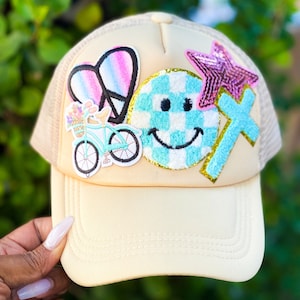 May include: A beige trucker hat with a variety of colorful patches, including a peace sign, a smiley face, a star, and a cross.