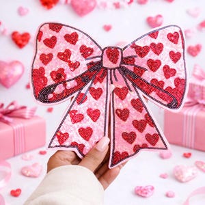 May include: A large, pink sequined bow decorated with red heart shapes. The bow is held in a hand, with a background of pink hearts and gift boxes. The bow's edges are outlined in black sequins, adding contrast to the design. A festive decoration for Valentine's Day.