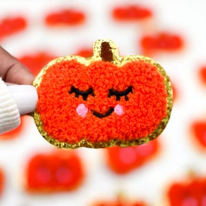 May include: A fuzzy orange pumpkin patch iron-on patch with a smiling face, closed eyes, and pink cheeks. The patch has a gold glitter outline.