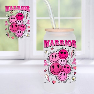 May include: A frosted glass tumbler with a pink and white design. The tumbler has a wooden lid and a glass straw. The design features the word "WARRIOR" in pink letters with a pink ribbon and smiley faces. The words "Believe", "Strength", "Courage", "Love", "Hope", "Faith", "Family", "Support", "Awareness", and "Fight" are also included in the design.