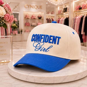 May include: A baseball cap with a cream-colored crown and a bright blue brim. The front of the cap features the text CONFIDENT Girl embroidered in bold blue lettering. The cap has a small blue button at the top center and blue eyelets on the sides.