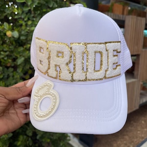 May include: White trucker hat with the word "BRIDE" in gold glitter letters. The hat features a white mesh back and a white front with a decorative ring patch. A stylish accessory for a bridal event.