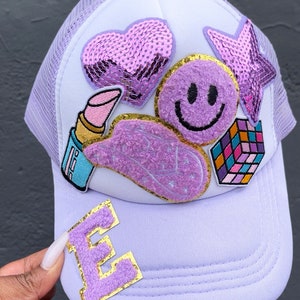May include: Purple and white trucker hat with a variety of colorful patches, including a heart, a smiley face, a star, a lipstick, and a Rubik's cube. The hat also has a purple felt letter 'E' on the front.