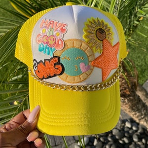 May include: Yellow trucker hat with a white crown featuring colorful embroidered patches that say "Have a Good Day" and "OMG". The hat has a gold chain trim.