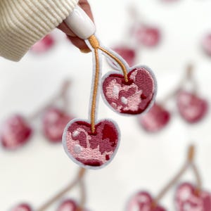 May include: Two pink and white embroidered cherries with gold stems. The cherries are on a white background.