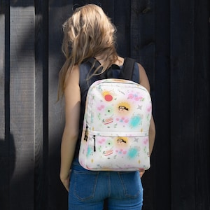 May include: A white backpack featuring a repeating pattern of anime-style character faces, bowls of ramen, blue popsicles, pink flowers, and shuriken stars. The bag includes a main compartment and a smaller front pocket, both with black zipper pulls.