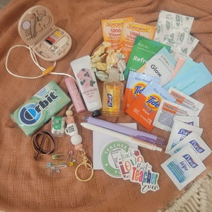 May include: A collection of small items, including a sewing kit, hand sanitizer, lip balm, gum, and Tide stain remover packets, arranged on a peach-colored fabric. There are also stickers that say "Teacher" and "Best Teacher Ever".