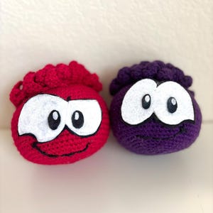 May include: Two crocheted plush toys, one red and one purple, each with white eyes and a smiling mouth. The red toy has a red, curly top, while the purple toy has a purple, curly top. The toys are round and have a cartoonish appearance.