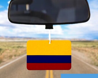 May include: A rectangular car air freshener with the flag of Colombia, featuring yellow, blue, and red horizontal stripes, hangs from a rearview mirror. The background shows a road and a blue sky.