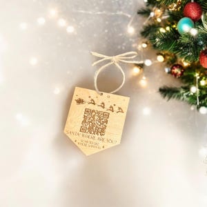 May include: Wooden Christmas ornament with a QR code and the text "SANTA! WHERE ARE YOU? SCAN TO SEE WHERE SANTA IS". The ornament features a laser-etched design of Santa's sleigh and reindeer. A string is attached for hanging.