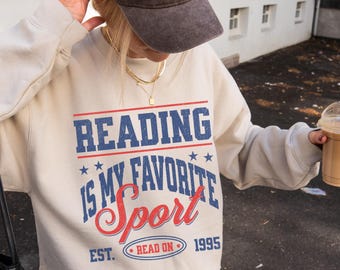 May include: A beige sweatshirt with the text "READING IS MY FAVORITE SPORT" in blue and red. The image also shows a brown baseball cap with "La Paris" text, a black purse, and a coffee drink.