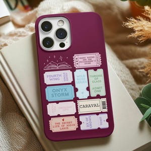 Può includere: Una custodia per telefono color borgogna con una varietà di adesivi colorati a forma di biglietto che presentano titoli di libri come "The Secret Life of Addie LaRue", "Six of Crows", "Caraval", "Throne of Glass", "Wild Rose Series", "Onyx Storm", "Fourth Wing" e "Harry Potter e la pietra filosofale".