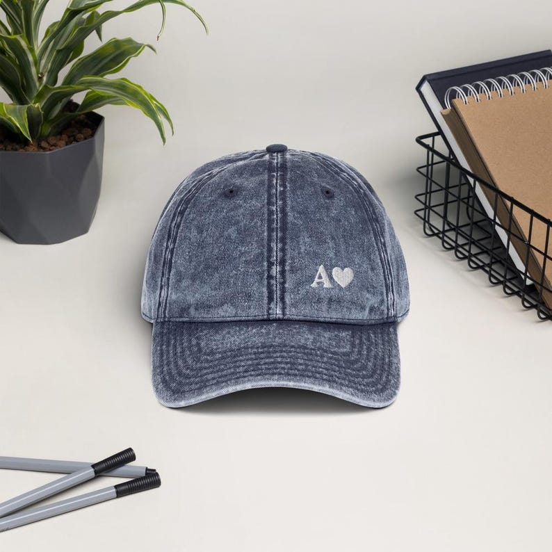 May include: A blue denim baseball cap with a white embroidered letter A and a small heart.