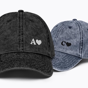 May include: Two denim baseball caps with embroidered letters and hearts. The black cap has a white "A" and heart, and the blue cap has a black "C" and heart.