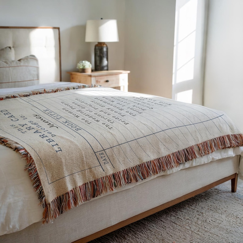 May include: A beige throw blanket with a library card design, featuring a list of book titles and the text "This Library Card belongs to: James." The blanket has a fringed edge with red, orange, and brown threads. It is placed on a bed.
