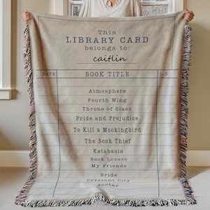 May include: A beige throw blanket with a library card design. The text reads "This LIBRARY CARD belongs to: caitlin" and lists book titles. The blanket has a fringed edge and is held up by a person.