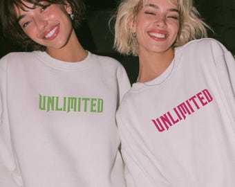 May include: Two people wearing white sweatshirts with the word "UNLIMITED" printed on the front. One sweatshirt has green text, and the other has pink text. The people are smiling.