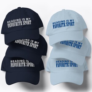 May include: A stack of navy blue and light blue baseball caps. Each cap has the text "READING IS MY FAVORITE SPORT" printed on the front in white or blue. The caps are made of a soft fabric and have a curved brim.