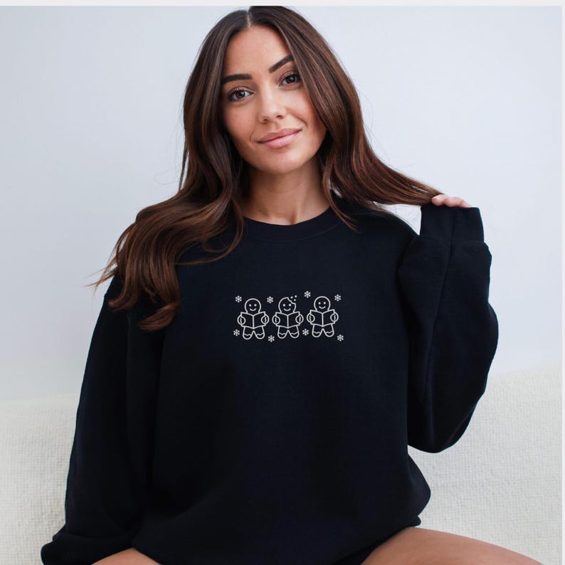 Pode incluir: Camisola preta com um desenho bordado branco de tr&ecirc;s bonecos de gengibre lendo livros, com flocos de neve. A camisola &eacute; de gola redonda e parece ser feita de um material macio.