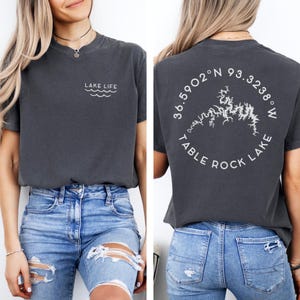 Puede incluir: Camiseta gris oscuro con el texto "LAKE LIFE" y un gráfico de olas en la parte delantera. La parte trasera de la camiseta presenta un diseño circular con el texto "TABLE ROCK LAKE" y coordenadas.