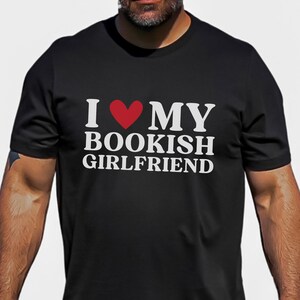 Puede incluir: Camiseta negra con texto blanco que dice "Amo a mi novia aficionada a los libros" con un corazón rojo.