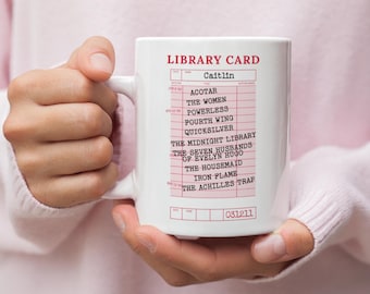 Puede incluir: Taza de cerámica blanca con un diseño de tarjeta de biblioteca rosa, con el nombre "Caitlin" y títulos de libros. La taza tiene un asa y se sostiene con las manos. El diseño incluye el número 031211.