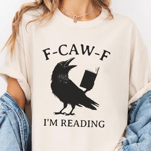 Pode incluir: Camiseta creme com um desenho de corvo preto segurando um livro, com o texto "F-CAW-F" acima e "I'M READING" abaixo. O corvo é retratado com o bico aberto. Uma peça de roupa casual.