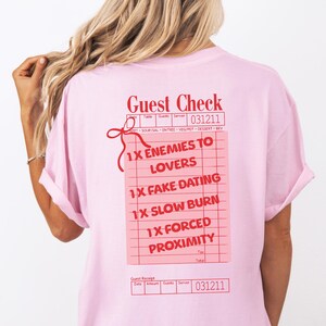 Pode incluir: Camiseta rosa com um gráfico vermelho e branco de um cheque de restaurante com o texto "1X Enemies to Lovers", "1X Fake Dating", "1X Slow Burn" e "1X Forced Proximity".