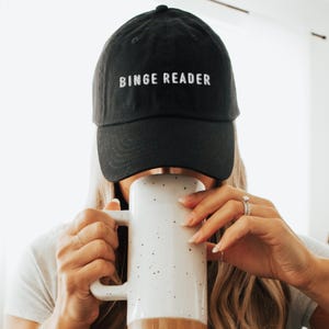 May include: Black baseball cap with the embroidered text "BINGE READER". The cap is worn by a person holding a large white mug with brown accents and black speckles. The person is wearing a ring on their finger.