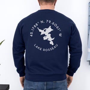 May include: Navy blue sweatshirt with a white graphic of Lake Rosseau, including coordinates and the lake's name. The design is circular and centered on the back of the sweatshirt. The sweatshirt is a solid color.