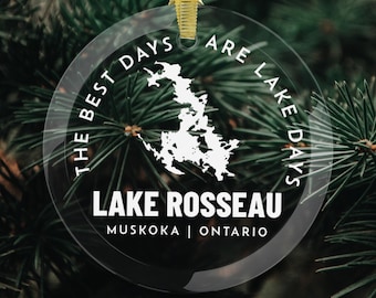 May include: A clear glass Christmas bauble with a gold ribbon. The bauble features a white map of Lake Rosseau, Ontario, with the text "THE BEST DAYS ARE LAKE DAYS" and "LAKE ROSSEAU MUSKOKA | ONTARIO". The bauble hangs against a backdrop of green pine needles.