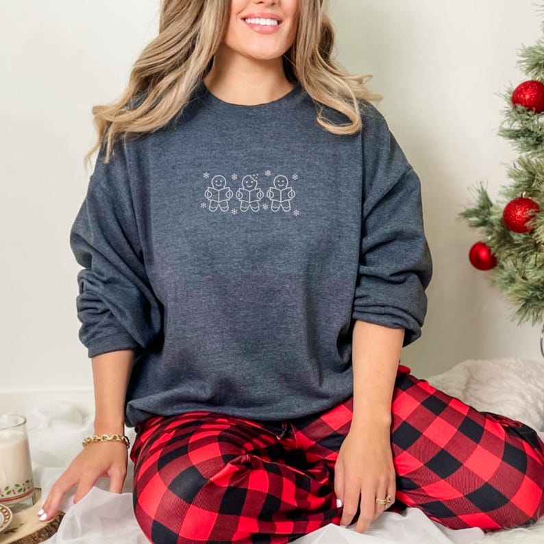 Pode incluir: Um moletom cinza escuro com um desenho bordado branco de tr&ecirc;s bonecos de gengibre e flocos de neve. A pessoa est&aacute; usando cal&ccedil;as de pijama xadrez vermelhas e pretas. Uma pequena &aacute;rvore de Natal com enfeites vermelhos est&aacute; ao fundo.