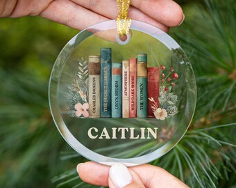 May include: A clear, round Christmas ornament with a gold ribbon. The ornament features a stack of vintage books with titles like "Charles Dickens" and "The Secret Garden." The name "CAITLIN" is printed below the books. Floral accents complete the design.