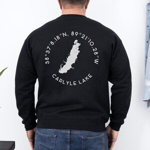 May include: Black sweatshirt featuring a white graphic of Carlyle Lake with coordinates and the lake's name. The design is circular, with the lake's shape in the center. The sweatshirt is a solid black color.
