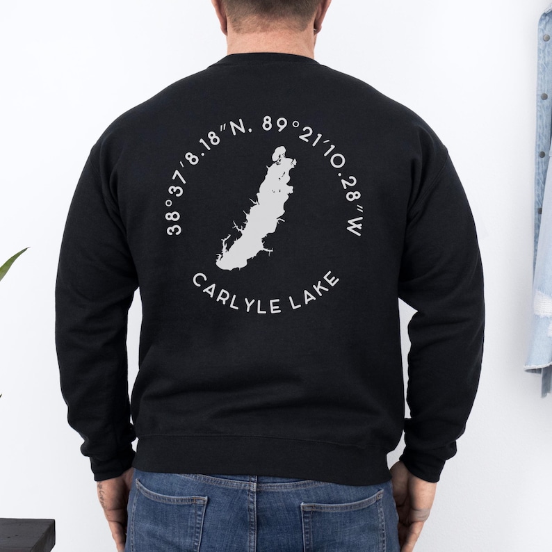 May include: Black sweatshirt featuring a white graphic of Carlyle Lake with coordinates and the lake's name. The design is circular, with the lake's shape in the center. The sweatshirt is a solid black color.