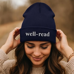 May include: A navy blue beanie hat with the white text "well-read" embroidered on the front. The hat is made of a soft, knitted material. The beanie is shown on a person.