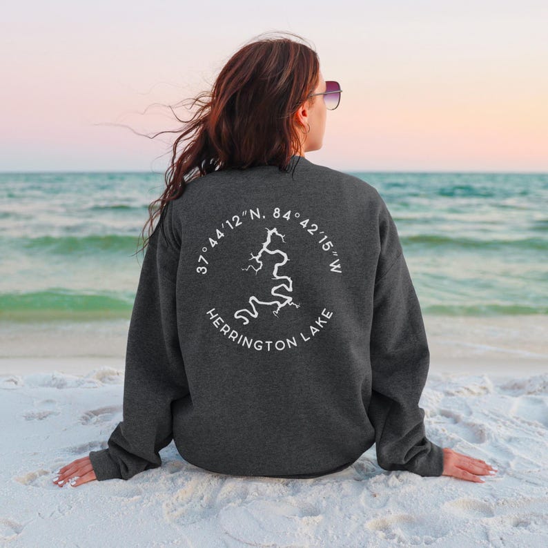 May include: Dark gray sweatshirt with a white graphic of a lake and the text "Herrington Lake" in a circle. The sweatshirt also features the coordinates 37&deg;44'12"N, 84&deg;42'15"W. The person is sitting on a sandy beach.